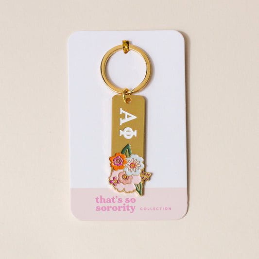 The Darling Effect’s Alpha Phi Enamel Keychain features Greek letters and floral designs on a gold keychain, displayed on a white card with a light pink bottom—perfect as a sorority gift from the That’s So Sorority collection.