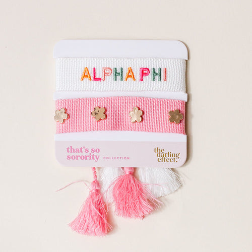The Darling Effect’s Alpha Phi Woven Bracelet Set includes two adjustable bracelets on a card—one white with colorful “ALPHA PHI” embroidery and one pink with gold flower studs—finished with pink and white tassels. Perfect for sisterhood gifting!.