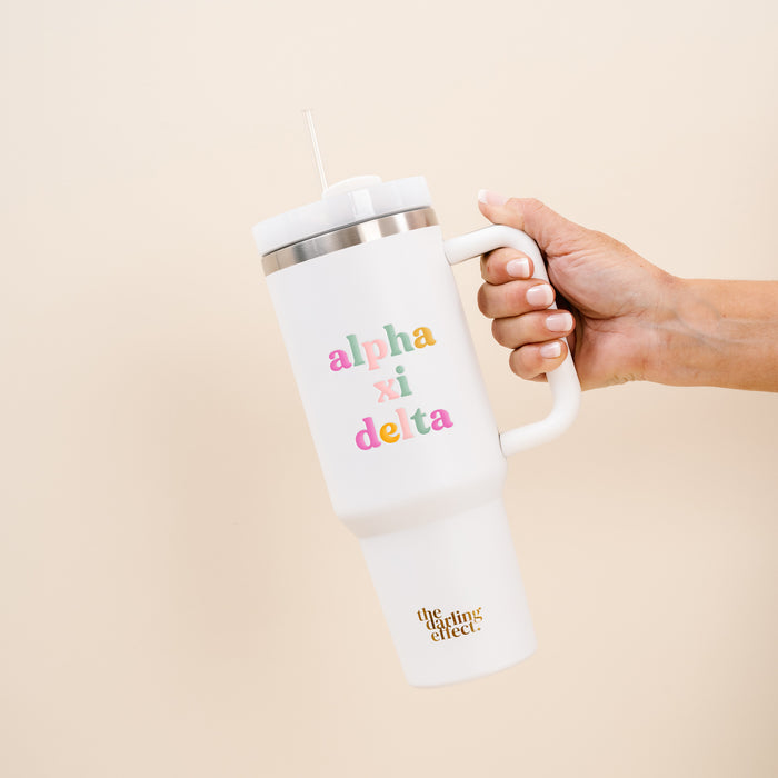 A hand holds The Darling Effect's Alpha Xi Delta 40 oz Take Me Everywhere White Tumbler, featuring a large handle, straw, colorful Alpha Xi Delta letters, and "the lifting effect" in gold near the bottom.