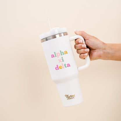 A hand holds The Darling Effect's Alpha Xi Delta 40 oz Take Me Everywhere White Tumbler, featuring a large handle, straw, colorful Alpha Xi Delta letters, and "the lifting effect" in gold near the bottom.