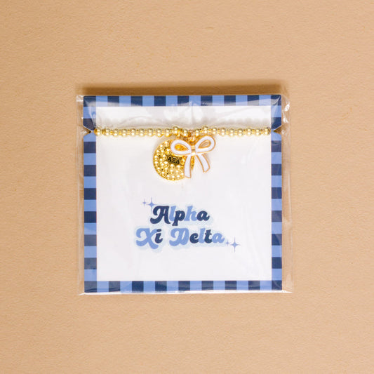 The Darling Effect's Alpha Xi Delta Bow Gold Charm Bracelet features gold beads, a crescent moon, and a white bow charm on a branded card with blue plaid—an ideal sorority jewelry gift for Alpha Xi Delta sisters.