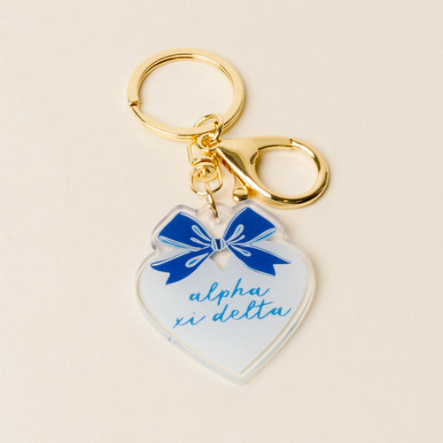 The Darling Effect Alpha Xi Delta Sweetheart Bow Acrylic Keychain features a heart shape, blue script, pastel bow, and gold keyring with clasp—perfect as a sisterhood gift.