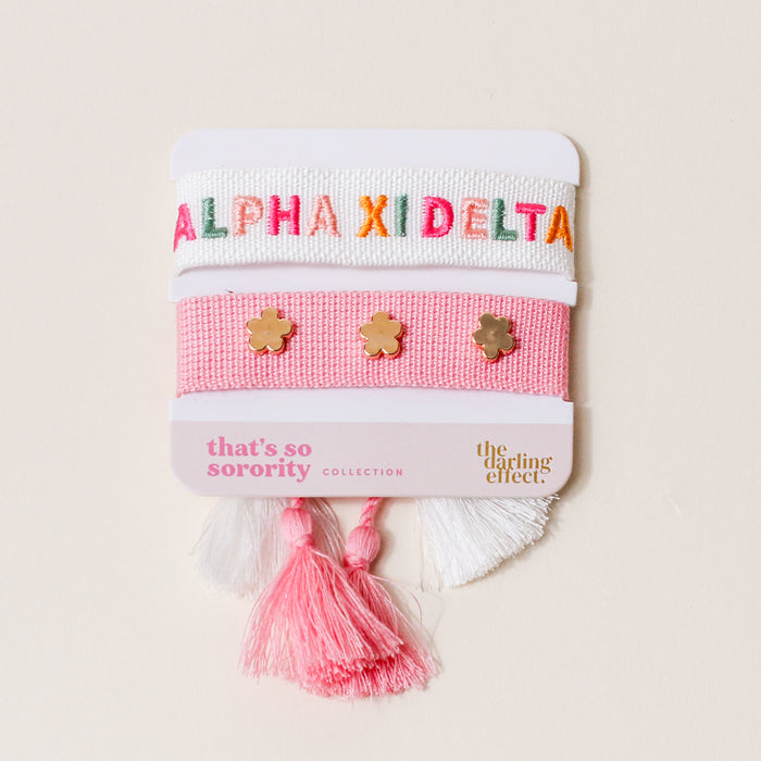 The Darling Effect’s Alpha Xi Delta Woven Bracelet Set includes two pastel pink and white adjustable bracelets—one with ALPHA XI DELTA in colorful letters, the other with three gold flower charms—making it a perfect sorority or sisterhood gift.