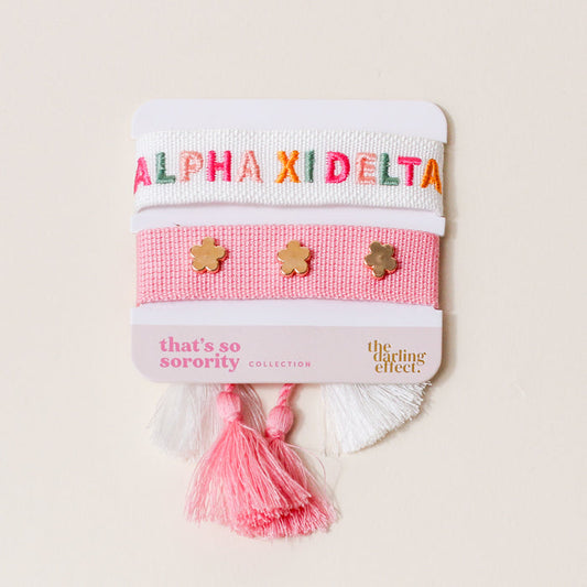 The Darling Effect’s Alpha Xi Delta Woven Bracelet Set includes two pastel pink and white adjustable bracelets—one with ALPHA XI DELTA in colorful letters, the other with three gold flower charms—making it a perfect sorority or sisterhood gift.