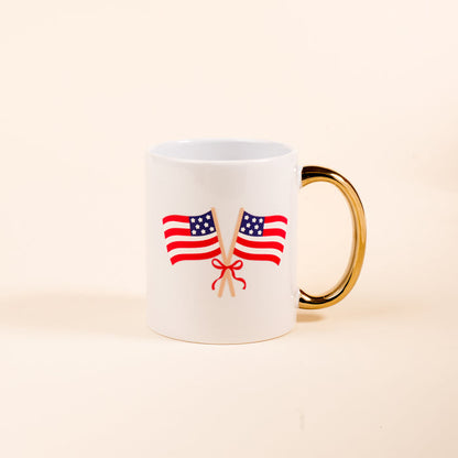 The Darling Effect's American Flags 11 oz Cozy Cute Ceramic Mug features two crossed American flags tied with a red ribbon and a golden handle, perfect for enjoying coffee or tea on a plain light background.