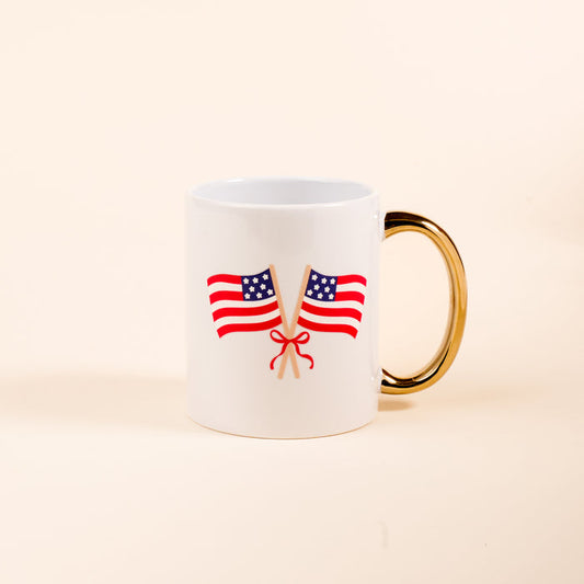 The Darling Effect's American Flags 11 oz Cozy Cute Ceramic Mug features two crossed American flags tied with a red ribbon and a golden handle, perfect for enjoying coffee or tea on a plain light background.