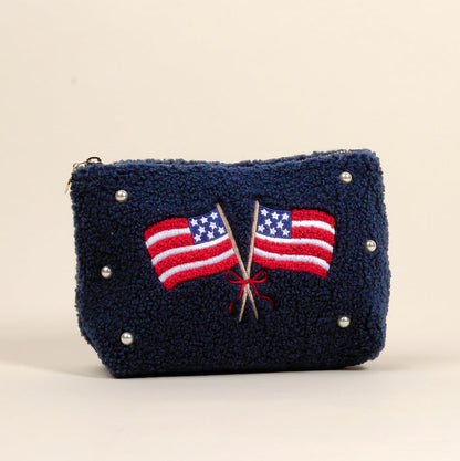 The Darling Effect’s American Flags Teddy Pouch is a blue textured sherpa zip pouch, accented with pearls and two crossed embroidered American flags tied with a red ribbon—an eye-catching accessory.