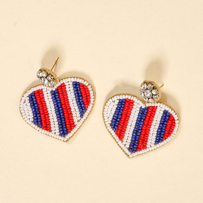 Americana Heart Beaded Earrings by The Darling Effect feature vibrant heart shapes with red, white, and blue bead stripes and a clear rhinestone cluster. These playful earrings are shown on a light beige background.