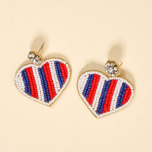 Americana Heart Beaded Earrings by The Darling Effect feature vibrant heart shapes with red, white, and blue bead stripes and a clear rhinestone cluster. These playful earrings are shown on a light beige background.