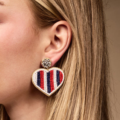 The Darling Effect’s Americana Heart Beaded Earrings feature hand-beaded red, white, and blue stripes with a gold trim, adding a playful touch to any look.