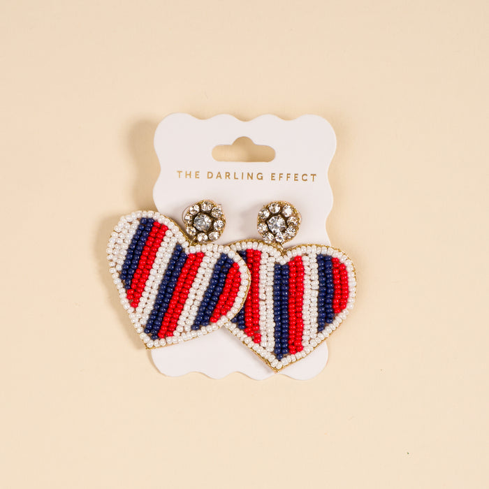 The Darling Effect's Americana Heart Beaded Earrings feature vibrant red, white, and blue stripes, heart shapes, and sparkly round studs. Each playful pair comes attached to a branded card.