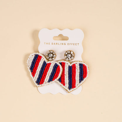 The Darling Effect's Americana Heart Beaded Earrings feature vibrant red, white, and blue stripes, heart shapes, and sparkly round studs. Each playful pair comes attached to a branded card.