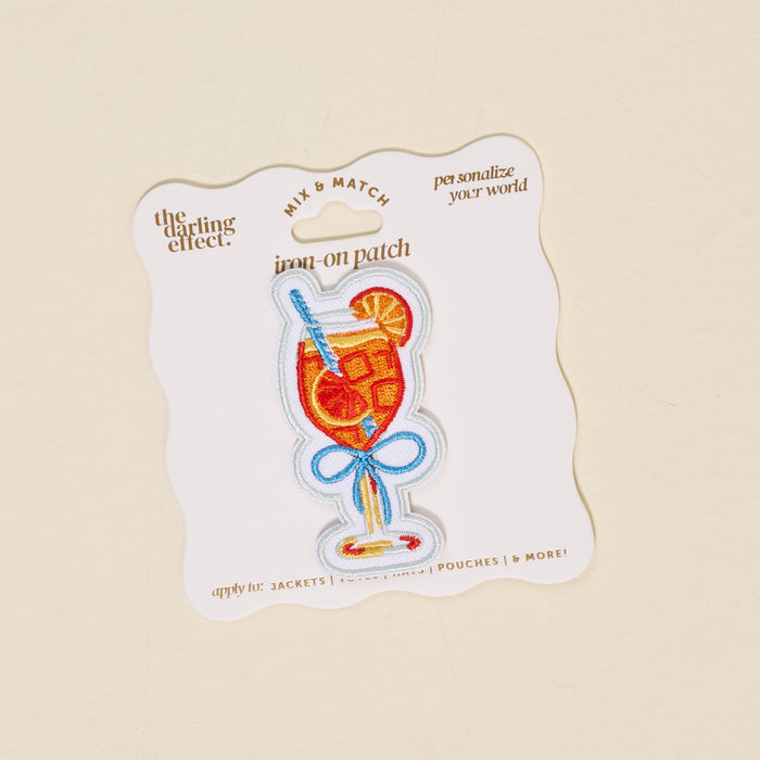 The Darling Effect’s Aperol Spritz Mix & Match Iron-On Patch features a colorful cocktail glass with garnishes, ice, and a blue straw—perfect for customizing your look. Comes on a white backing card with gold and tan text.