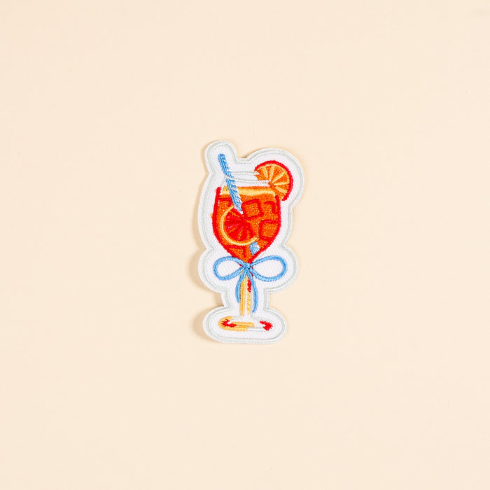 The Darling Effect's Aperol Spritz Mix & Match Iron-On Patch features a glass with a red drink, ice cubes, straw, lemon slice, and blue bow—perfect for customizing bags or jackets. Set on a plain beige background.