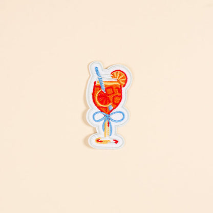 The Darling Effect's Aperol Spritz Mix & Match Iron-On Patch features a glass with a red drink, ice cubes, straw, lemon slice, and blue bow—perfect for customizing bags or jackets. Set on a plain beige background.