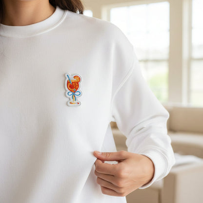 Someone stands in a bright, softly lit room wearing a white sweatshirt featuring The Darling Effect's Aperol Spritz Mix & Match Iron-On Patch—a colorful cocktail glass with fruit and a straw—on the chest.