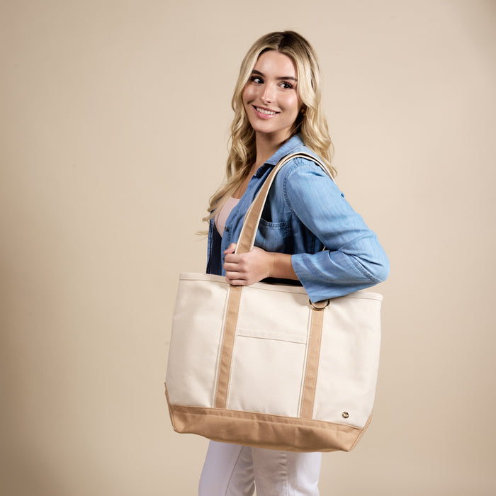 A woman with long blonde hair, in a light blue denim shirt and white pants, smiles while holding The Darling Effect’s Beige The Signature Canvas Tote Bag over her shoulder against a plain beige background.