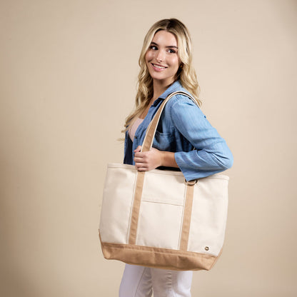 A woman with long blonde hair, in a light blue denim shirt and white pants, smiles while holding The Darling Effect’s Beige The Signature Canvas Tote Bag over her shoulder against a plain beige background.