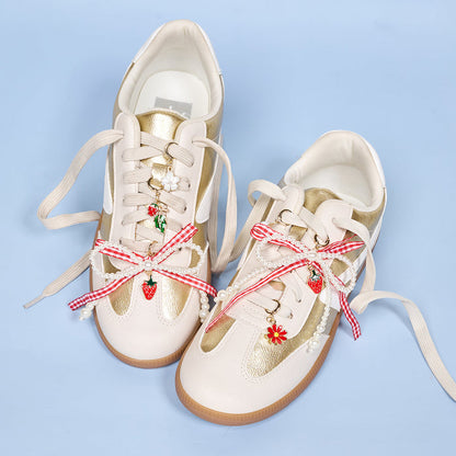 White and gold sneakers with beige laces are decorated using The Darling Effect’s Berry Fresh Dazzle Shoe Charm Pack—featuring red and white gingham bows, strawberries, flowers, and pearl charms—set against a light blue background.