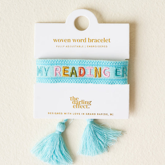 The Darling Effect's Best Weekend Woven Bracelet, crafted from lightweight blue material with MY READING ERA embroidered in multicolors, comes carded with a tassel detail below and The Darling Effect logo displayed above.
