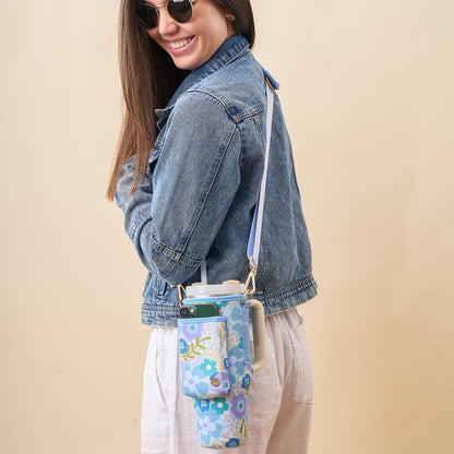 A smiling woman in sunglasses and a denim jacket wears The Darling Effect’s Beyond Blooms Blue Green Tumbler Sling crossbody over white pants. The floral-patterned neoprene bag features an adjustable strap and phone pocket, perfect as a stylish tumbler holder.