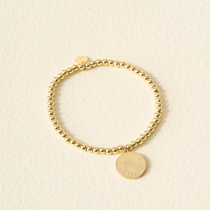 The Darling Effect's Birth Flower Beaded Charm Bracelet, featuring a round engraved pendant, rests on a light textured surface—ideal for adding a personalized touch to your jewelry collection.