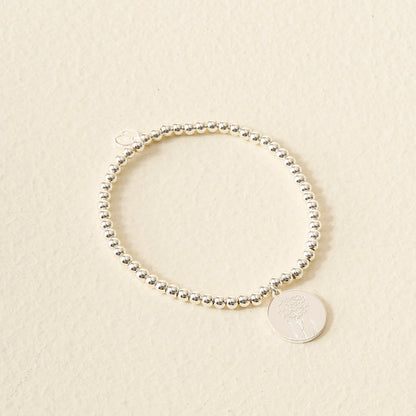 The Darling Effect Birth Flower Beaded Charm Bracelet, featuring a round floral-engraved charm, is displayed on a light beige textured surface.