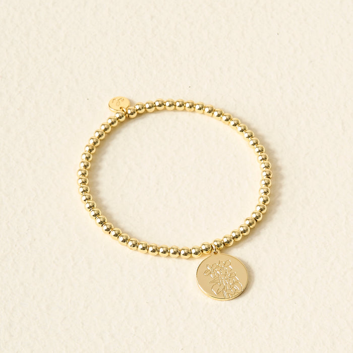 The Darling Effect Birth Flower Beaded Charm Bracelet features gold beads and a round charm with an engraved floral design—ideal as a personalized charm or birth flower bracelet.