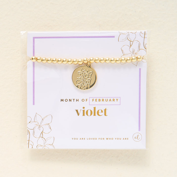 The Darling Effect Birth Flower Beaded Charm Bracelet for February features a gold beaded design with a round floral charm, displayed on a card with "You are loved for who you are," violet accents, and gold details—an ideal birth flower gift.