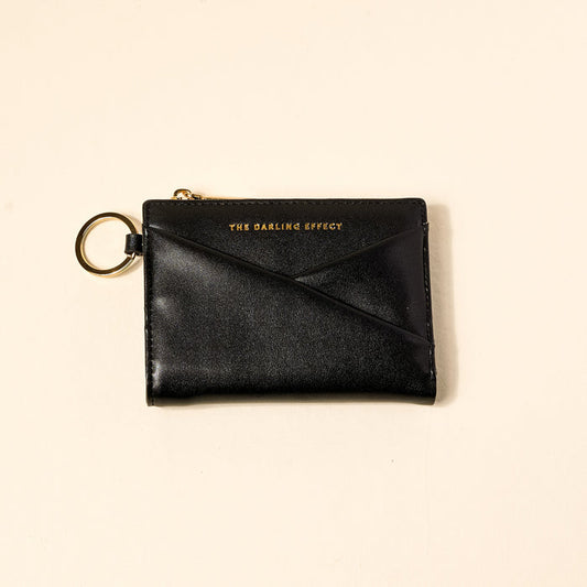 The Darling Effect’s Black Luxe Pocket Wallet, made from black vegan leather with a gold zipper and key ring, features the brand name embossed in gold on the front and is shown on a light beige background.
