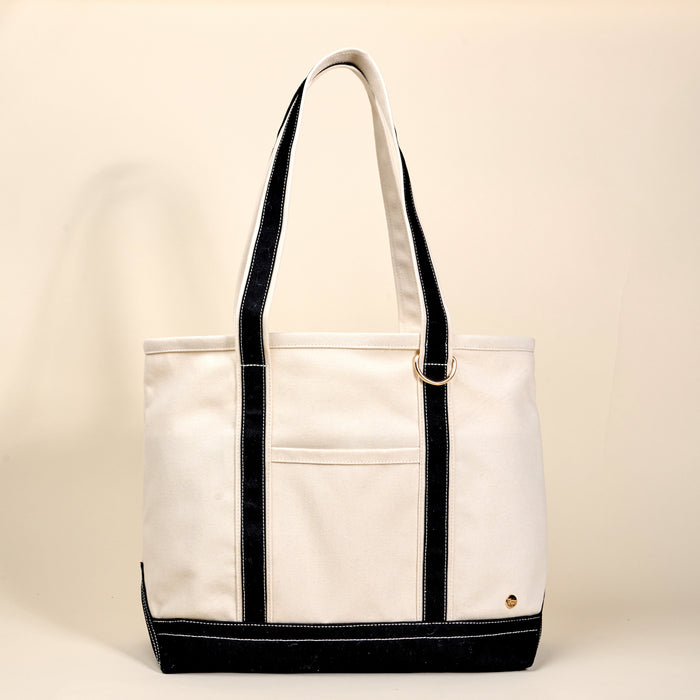 The Darling Effect's Black The Signature Canvas Tote Bag features durable black handles, a front pocket, and a gold ring accent. Designed for everyday use, it stands out against a plain light beige background.