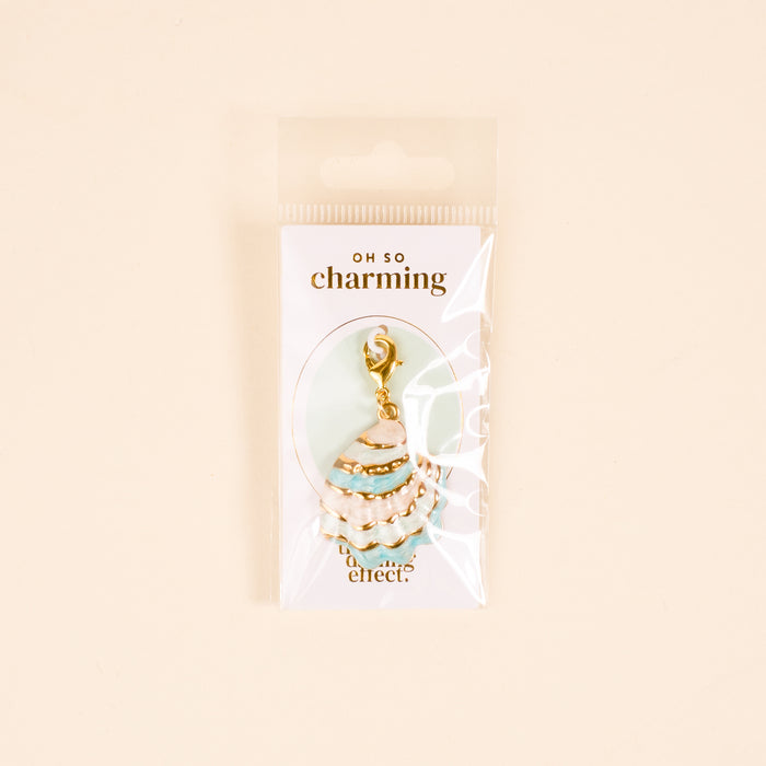 The Darling Effect’s Blue Shell Beauty Jewelry Charm Gold, shaped like a seashell with turquoise enamel, is shown in clear packaging labeled “Oh So Charming” against a pale background.