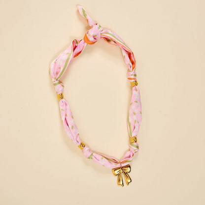 The Darling Effect's Blushing Hour Make a Statement Scarf Necklace features pink floral patterns, gold bow, and beads—an effortlessly bold accessory for any look.
