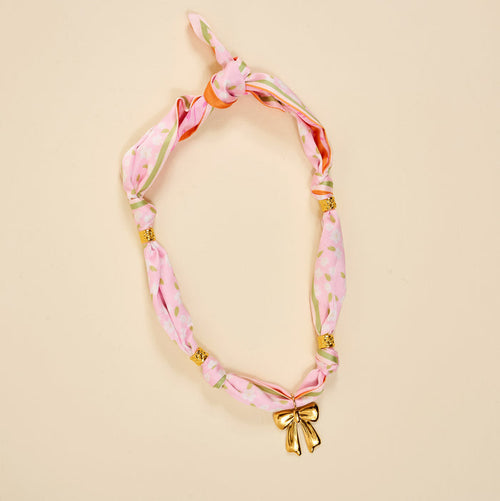 The Darling Effect's Blushing Hour Make a Statement Scarf Necklace features pink floral patterns, gold bow, and beads—an effortlessly bold accessory for any look.