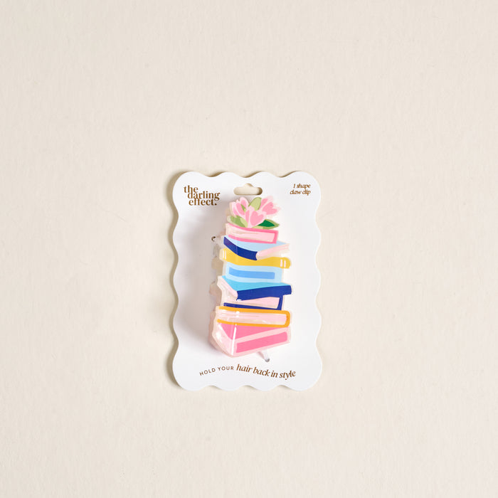 The Darling Effect's Book Nook Shape Up Claw Clip is a clear acrylic hair accessory shaped like stacked colorful books with pink flowers and green leaves, presented on a white backing card.