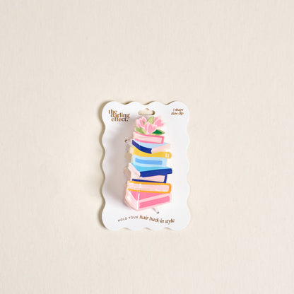 The Darling Effect's Book Nook Shape Up Claw Clip is a clear acrylic hair accessory shaped like stacked colorful books with pink flowers and green leaves, presented on a white backing card.