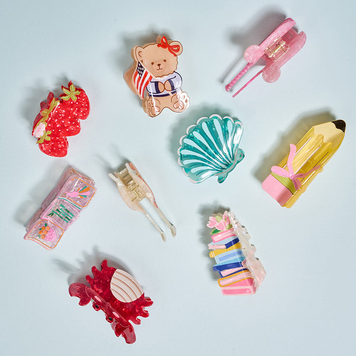Nine playful hair clips from The Darling Effect, including the Book Nook Shape Up Claw Clip, feature strawberries, a teddy bear, books, and abstract designs—perfect accessories for cute hairstyles on a light blue background.
