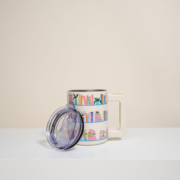 The Darling Effect’s Book Shelfie 14 oz Insulated Mug is a white mug with a large handle, featuring colorful book and plant illustrations. It comes with a clear plastic lid and is shown on a white surface against a neutral background.