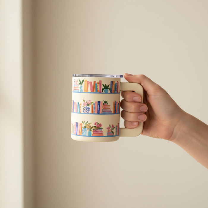 A hand holds The Darling Effect Book Shelfie 14 oz Insulated Mug, featuring colorful book and potted plant illustrations on shelves, set against a plain beige background.