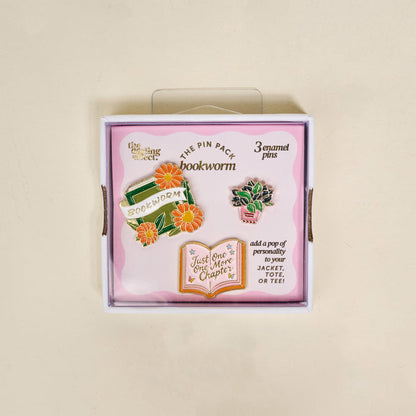 Bookworm The Pin Pack by The Darling Effect includes three collectible pins—a book with flowers, "Just One More Chapter" sign, and a potted plant—all packaged in a light pink box.