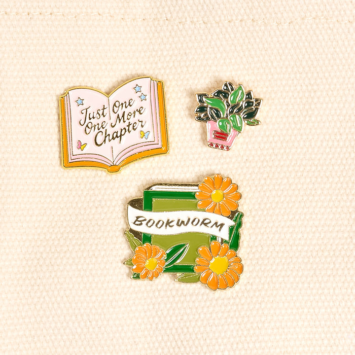Bookworm The Pin Pack by The Darling Effect features three enamel pins on beige: an open book with "Just One More Chapter," a potted green plant, and stacked books with yellow flowers and a Bookworm banner—perfect for book lovers!.
