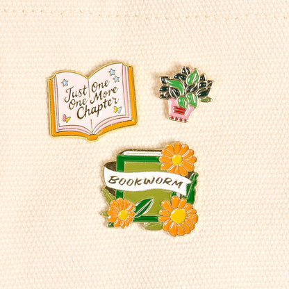 Bookworm The Pin Pack by The Darling Effect features three enamel pins on beige: an open book with "Just One More Chapter," a potted green plant, and stacked books with yellow flowers and a Bookworm banner—perfect for book lovers!.
