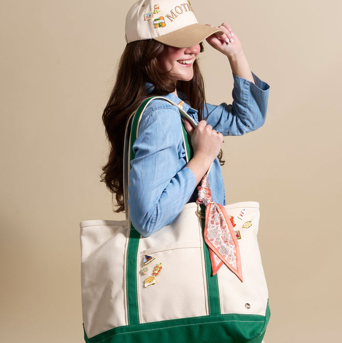 A young woman smiles as she adjusts her MOTHER baseball cap. She carries a canvas tote accented with The Darling Effect’s Bookworm The Pin Pack—featuring collectible pins—and a floral scarf tied to the handle.
