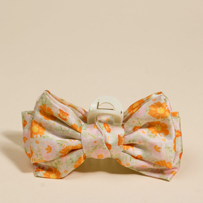 The Darling Effect’s Bouquet Beauty Sage Satin Bow Claw Clip features a large, floral-patterned bow in pink, white, and orange—an ideal accessory to elevate any updo. Shown against a plain beige background.