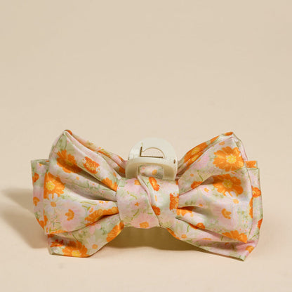 The Darling Effect’s Bouquet Beauty Sage Satin Bow Claw Clip features a large, floral-patterned bow in pink, white, and orange—an ideal accessory to elevate any updo. Shown against a plain beige background.