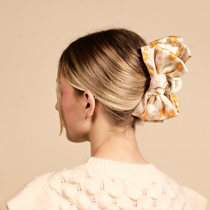 A woman with light brown hair in a low updo, secured with The Darling Effect's Bouquet Beauty Sage Satin Bow Claw Clip, is shown from the back wearing a cream textured sweater against a beige background.