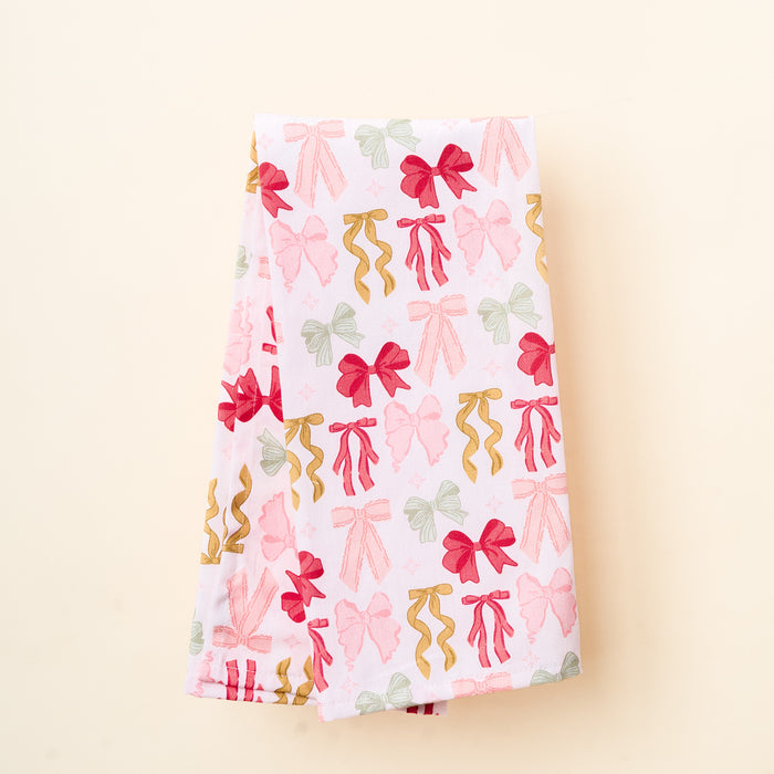 The Darling Effect's Bow Affair Tea Towel features a white fabric adorned with red, pink, gold, and green bow patterns, displayed neatly folded on a light beige background.
