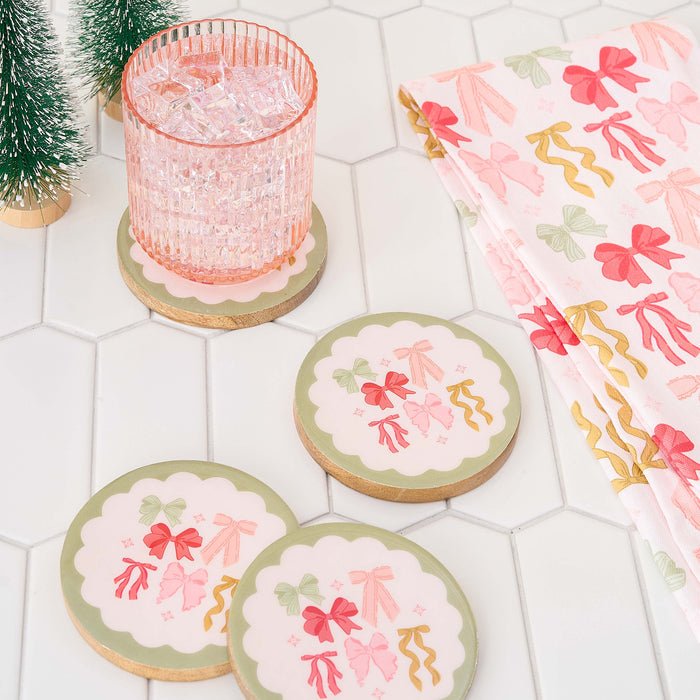 A pink glass with ice sits on a round coaster featuring colorful bows, with two similar coasters and The Darling Effect’s Bow Affair Tea Towel nearby, all arranged on white hexagonal tiles with small green trees in the corner.