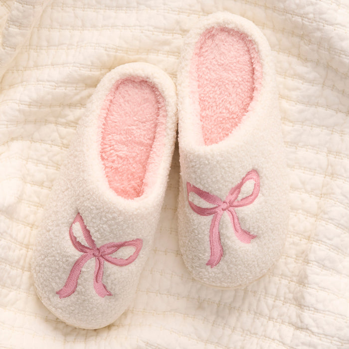The Bow Pink Fuzzy Slippers by the-darling-effect are cozy white slip-on slippers with pink interiors and embroidered bows, shown on a textured cream blanket.