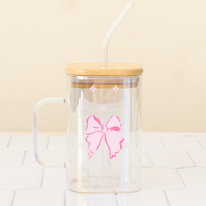 The Darling Effect’s Bow Square Glass Cup, a clear iridescent glass with handle, pink bow design, bamboo lid, and bent straw, sits on a white tiled surface against a light beige background.